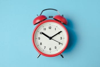 Photo of a traditional analog red alarm clock with red bells and white face with black numbers and mark, showing approximately 10:10 and 18 seconds, on light blue background.